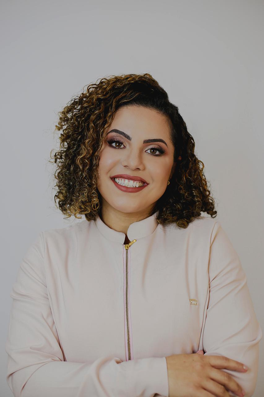 Dra. Priscila Barroso sorrindo, de jaqueta rosa clara e braços cruzados